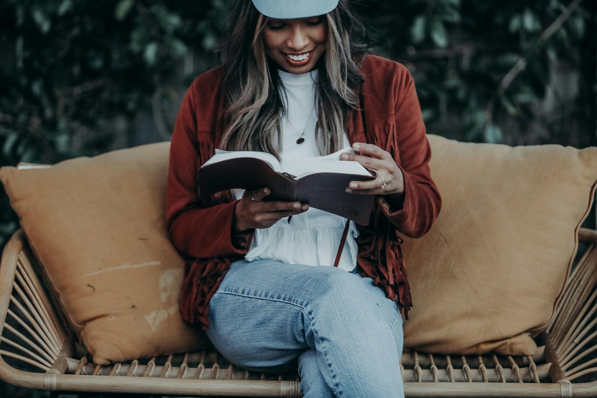A woman sitting on a couch, smiling as she reads an open Bible