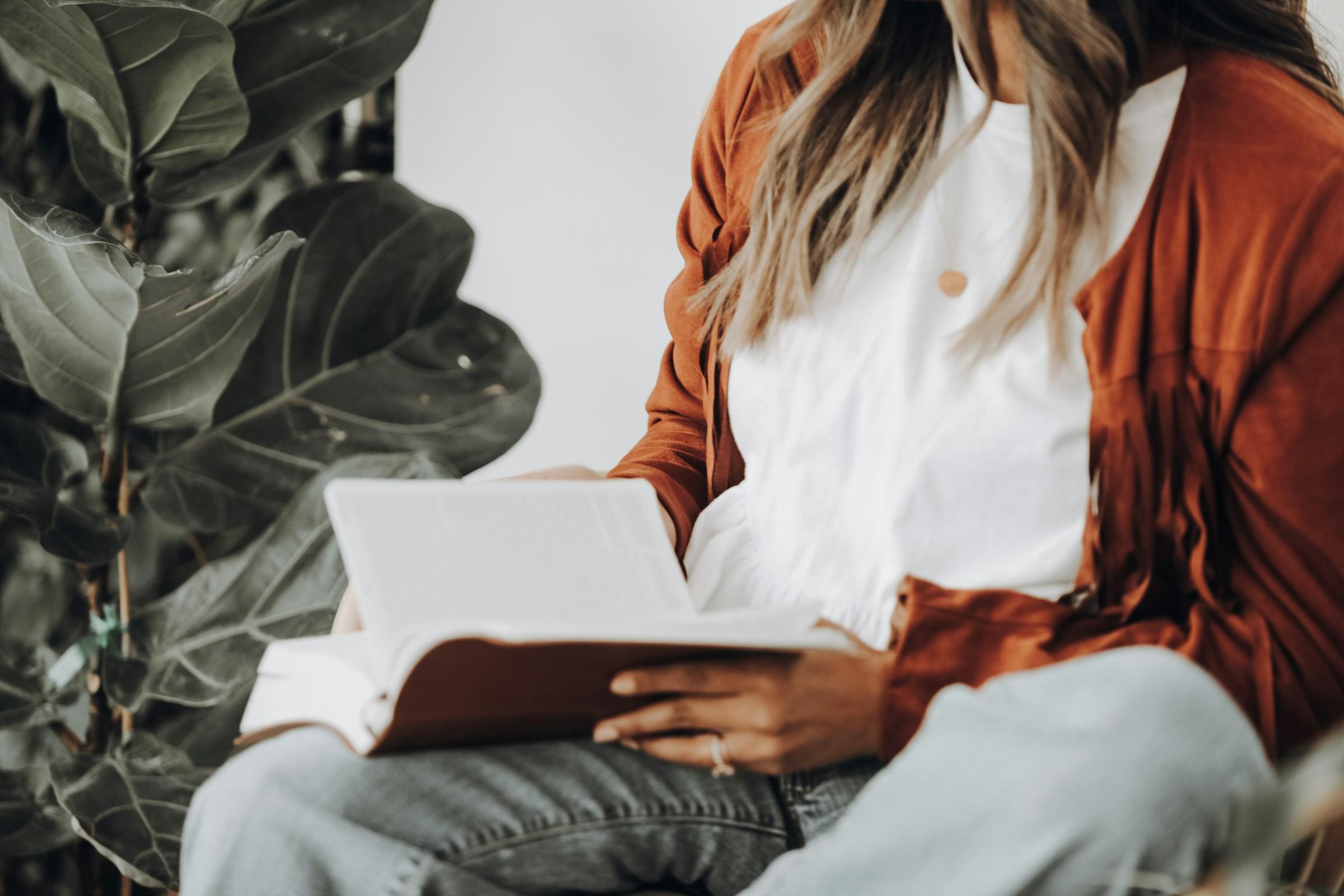 A woman in a rust-coloured jacket reading an open Bible beside a fiddle-leaf fig